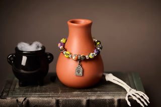 Halloween beaded bracelet draped on a terracotta vase with a black cauldron and skeleton hand prop