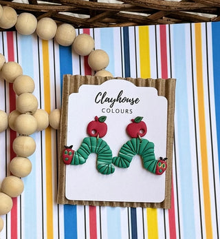 Handmade polymer clay earrings with red apple tops, green leaves, and textured green caterpillar dangles with red faces